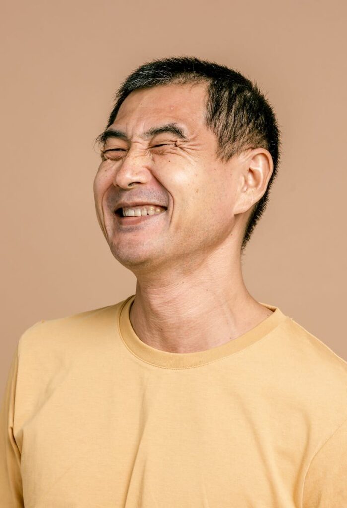 Portrait of a happy elderly man smiling with eyes closed, wearing a yellow t-shirt against a brown background.