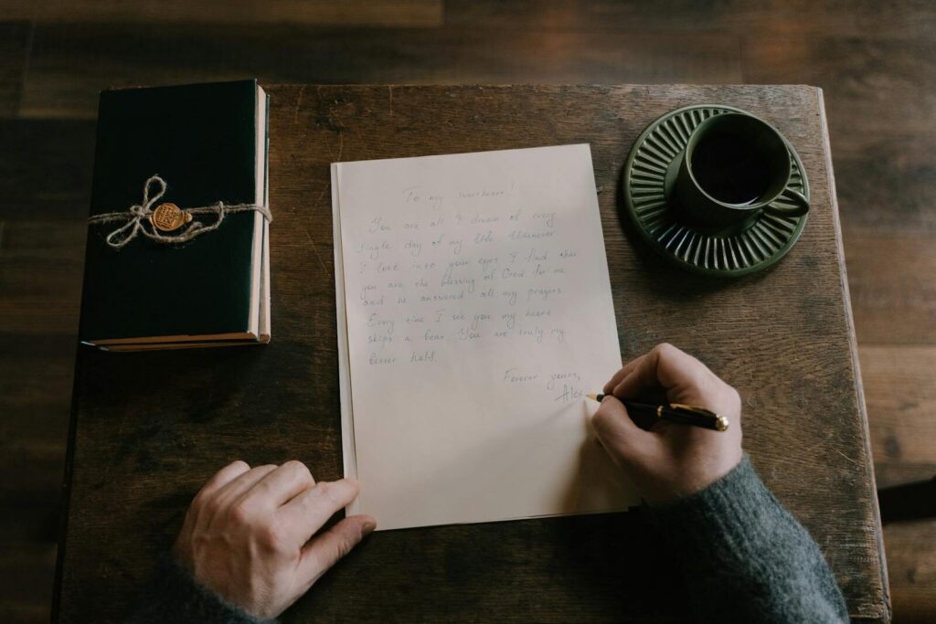 A person handwrites a letter alongside a journal and coffee cup on a wooden table.